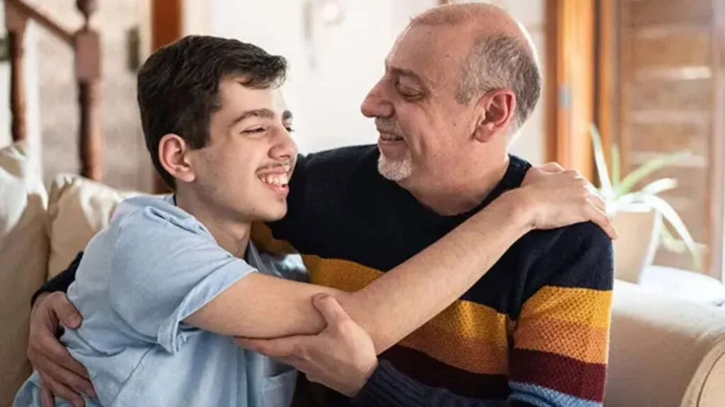 Smiling teenage boy with autism embracing his older father in a heartfelt hug, sitting in a cozy home setting