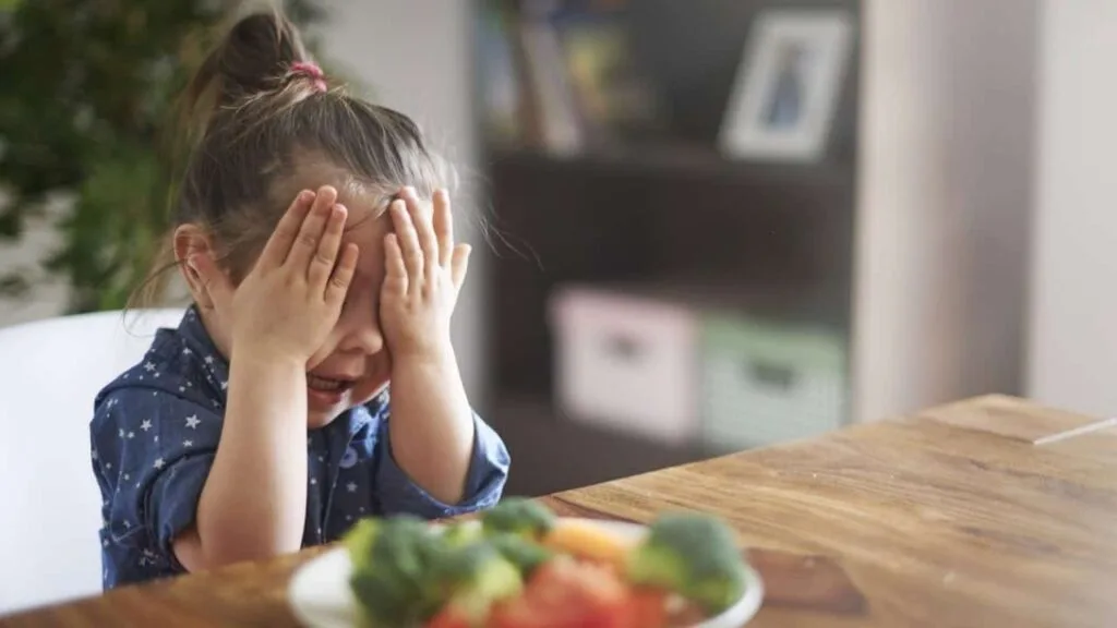 Young girl with pigtails crying and covering her face with her hands at a dining table, with a plate of vegetables in front of her