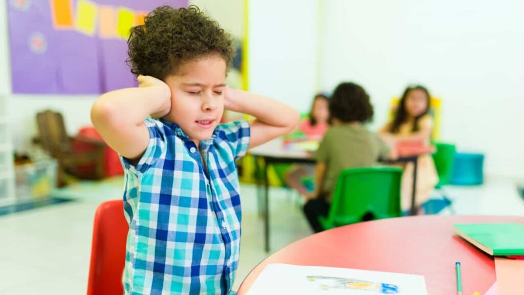 Young boy with curly hair covering his ears in a colorful classroom, appearing distressed by noise, with other children in the background.